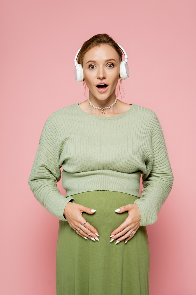 Embarazada mujer embarazada en traje verde tocando el vientre y el uso de auriculares aislados en rosa  - Foto, imagen