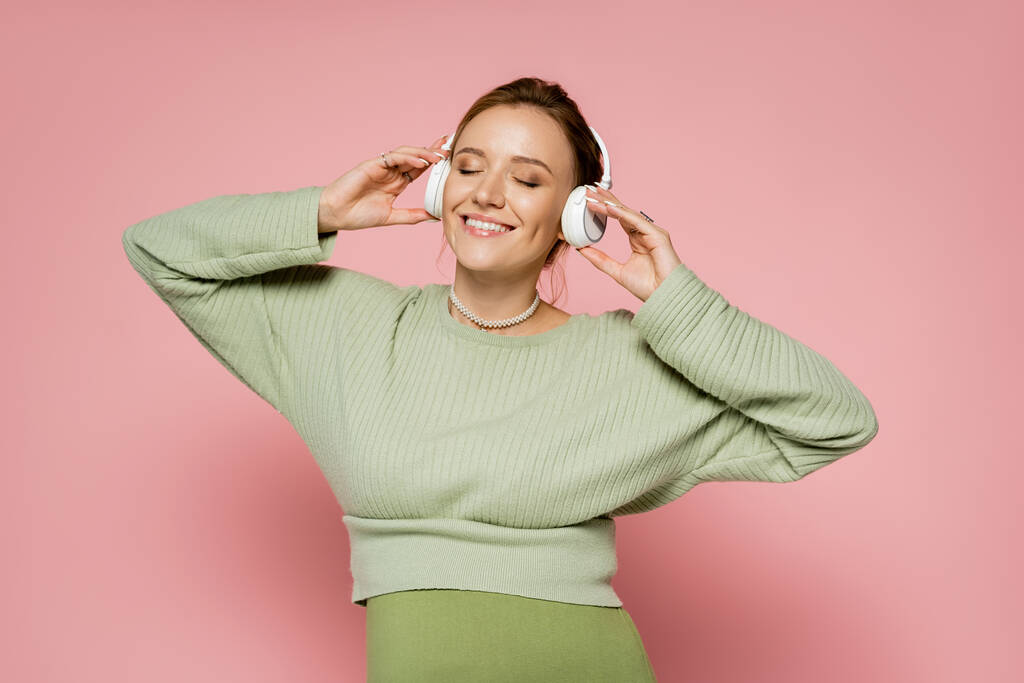 Positive pregnant woman closing eyes while listening music in headphones on pink background  - Photo, Image