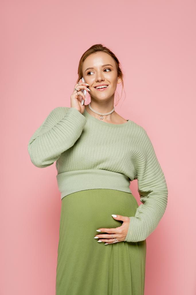Yeşil elbiseli, gülümseyen hamile bir kadın. Pembelerden izole bir şekilde akıllı telefondan konuşuyor.  - Fotoğraf, Görsel
