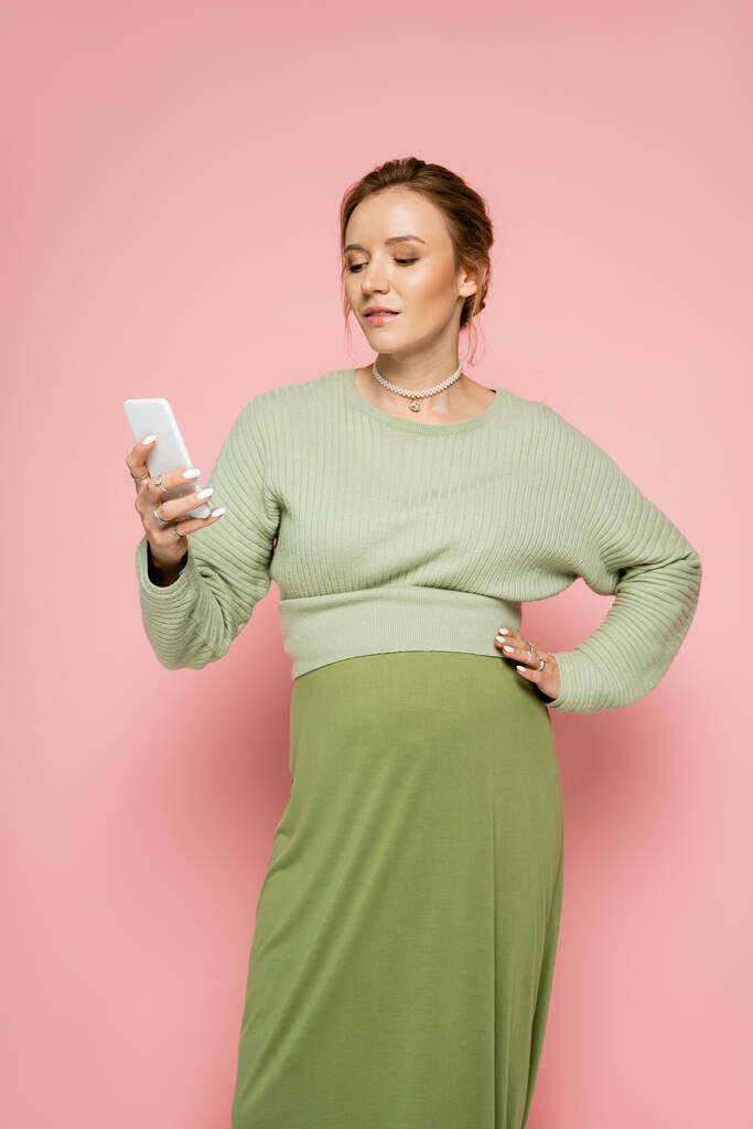 Junge schwangere Frau im grünen Outfit mit Smartphone und Händchen an der Hüfte auf rosa Hintergrund  - Foto, Bild