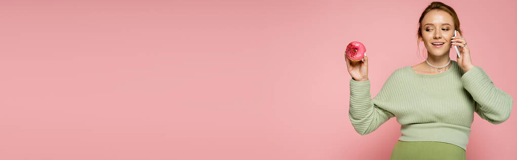 Счастливая беременная женщина говорит на смартфоне и глядя на сладкий пончик изолирован на розовый, баннер  - Фото, изображение