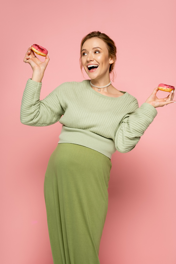 Mujer embarazada emocionada en suéter con rosquillas dulces sobre fondo rosa  - Foto, Imagen