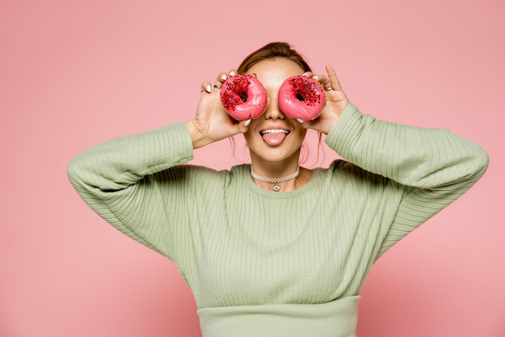 Jovem mulher saindo da língua e segurando donuts perto de olhos isolados em rosa  - Foto, Imagem