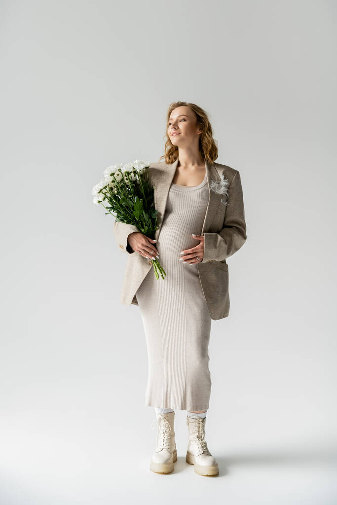 Gri arka planda beyaz çiçekler tutan, ceketli ve şık hamile bir kadın.  - Fotoğraf, Görsel