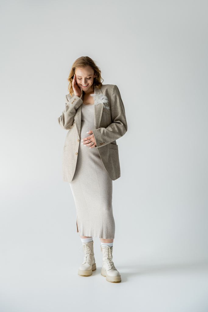 Full length of trendy pregnant woman in dress and boots standing on grey background  - Photo, Image