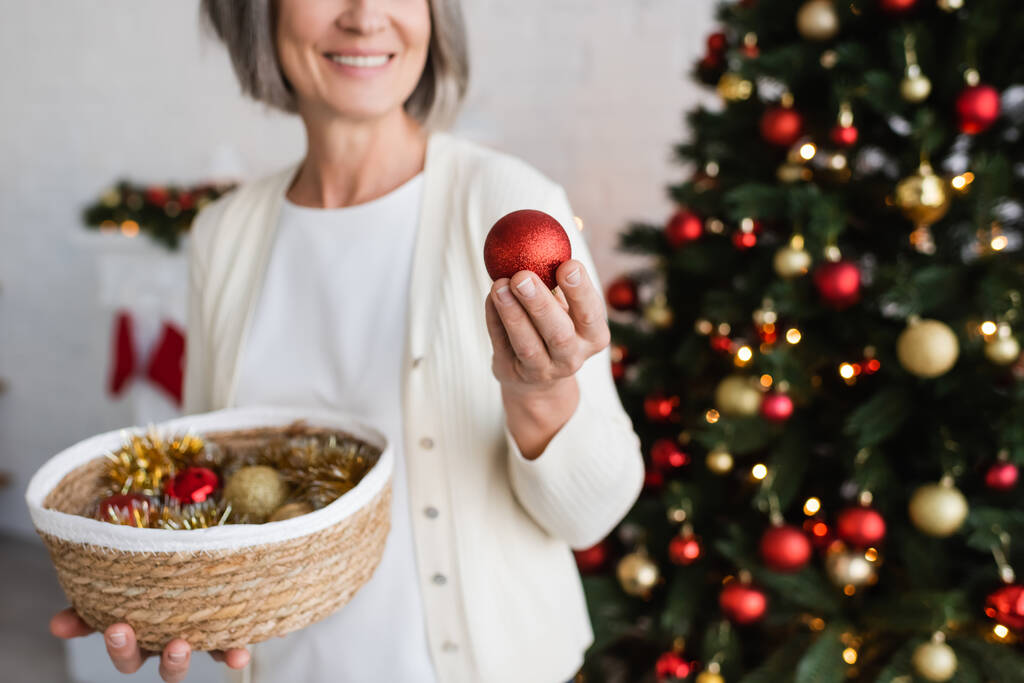 обрезанный вид веселой женщины средних лет с седыми волосами, держащей плетеную корзину и безделушку возле рождественской елки  - Фото, изображение