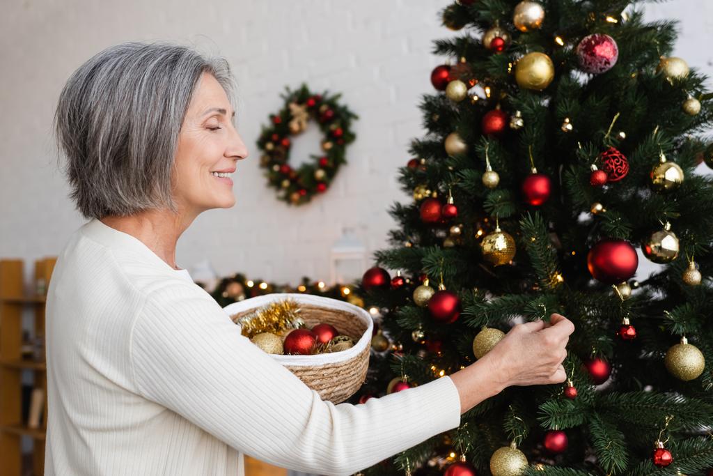 mujer de mediana edad feliz con pelo gris sosteniendo canasta de mimbre y decorando el árbol de Navidad  - Foto, Imagen
