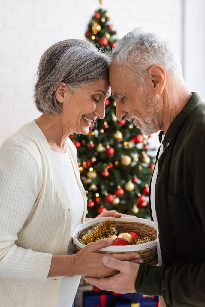 usmívající se pár středního věku se zavřenýma očima drží proutěný koš v blízkosti zdobené vánoční stromeček - Fotografie, Obrázek