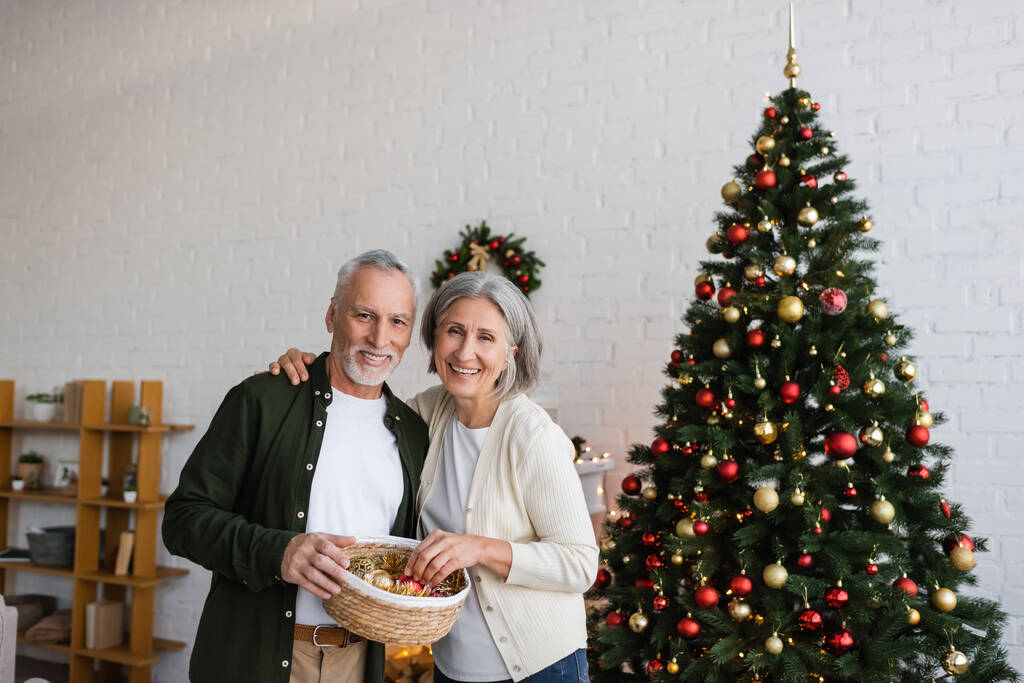 Gülümseyen orta yaşlı çift ellerinde sepet ve süslemeli noel ağacının yanında takılar tutuyorlar. - Fotoğraf, Görsel