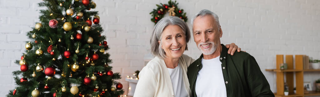 щаслива пара середнього віку, посміхаючись біля прикрашеної ялинки, банер
 - Фото, зображення