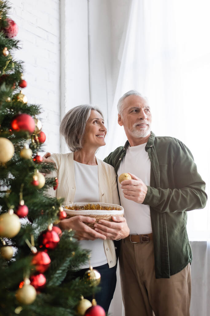 fröhlicher Mann mittleren Alters umarmt Ehefrau und schaut in der Nähe des geschmückten Weihnachtsbaums weg - Foto, Bild