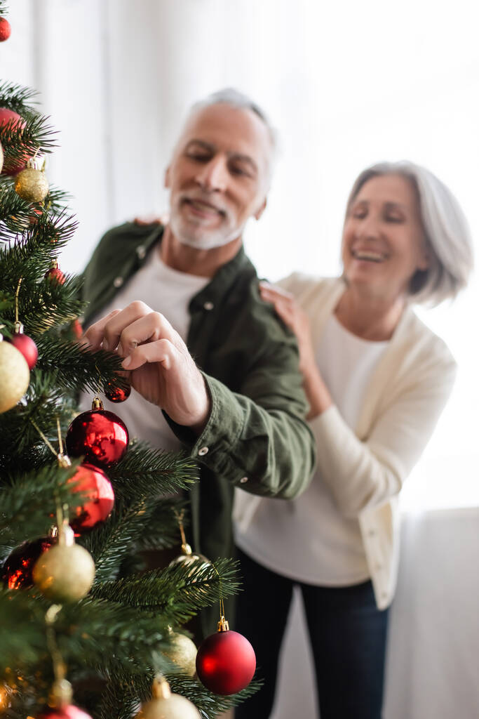 χαρούμενη μεσήλικη γυναίκα αγκαλιάζει θολή σύζυγο διακοσμώντας χριστουγεννιάτικο δέντρο - Φωτογραφία, εικόνα