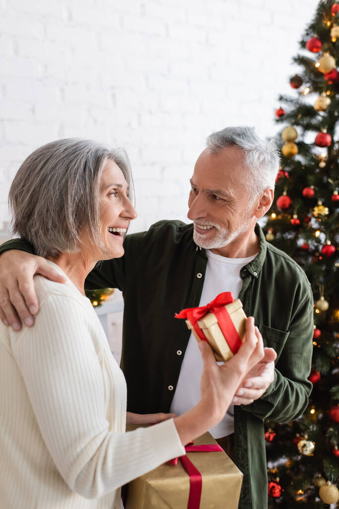 Улыбающаяся зрелая женщина с рождественским подарком рядом с веселым мужем и украшенной елкой  - Фото, изображение
