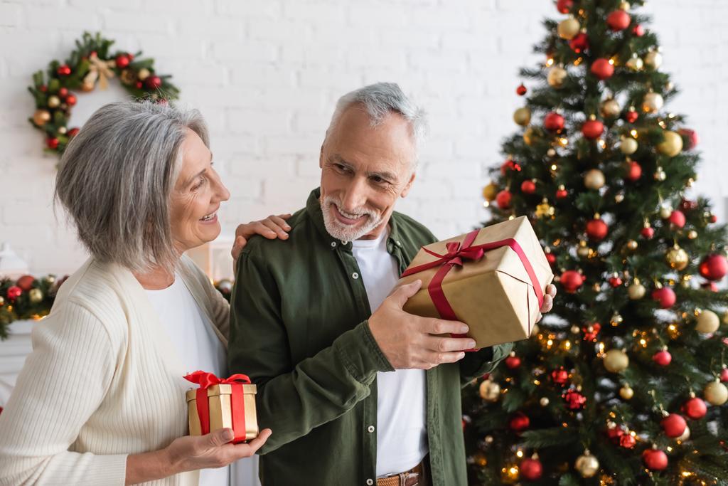 glimlachende volwassen vrouw met kerstcadeau en kijken naar vrolijke echtgenoot in de buurt versierde dennen - Foto, afbeelding