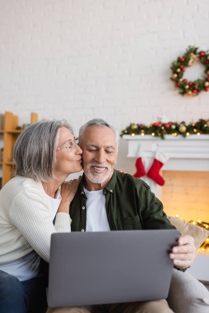 sonriente mujer de mediana edad besar mejilla de marido durante tener videollamada en el portátil en el día de Navidad - Foto, Imagen