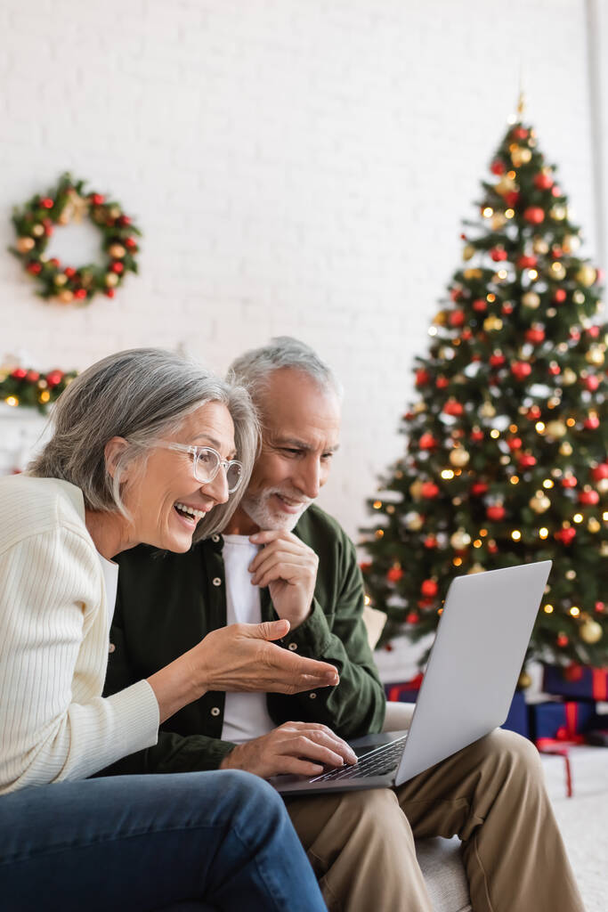 χαμογελαστή μεσήλικη γυναίκα να κάνει χειρονομίες κοντά στον σύζυγο και να έχει βιντεοκλήση στο φορητό υπολογιστή κατά τη διάρκεια των Χριστουγέννων  - Φωτογραφία, εικόνα