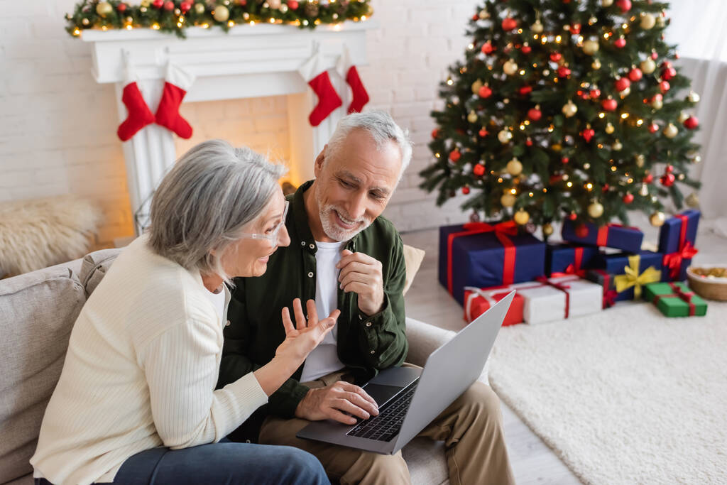 glimlachende vrouw van middelbare leeftijd wijzend naar gelukkige echtgenoot en het hebben van video call op laptop tijdens kerst  - Foto, afbeelding