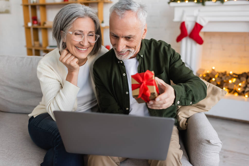 ευτυχισμένος μεσήλικας άνδρας κρατώντας χριστουγεννιάτικο δώρο κοντά στη γυναίκα και το laptop κατά τη διάρκεια της βιντεοκλήσης  - Φωτογραφία, εικόνα