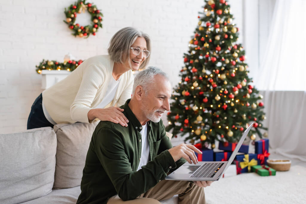alegre mujer de mediana edad en gafas abrazando marido durante la videollamada cerca de árbol de Navidad - Foto, Imagen
