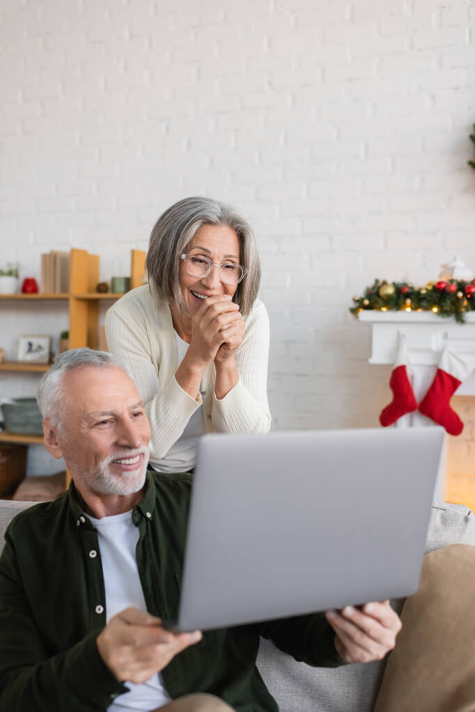 happy middle aged man showing laptop to flattered wife in glasses on christmas day  - 写真・画像