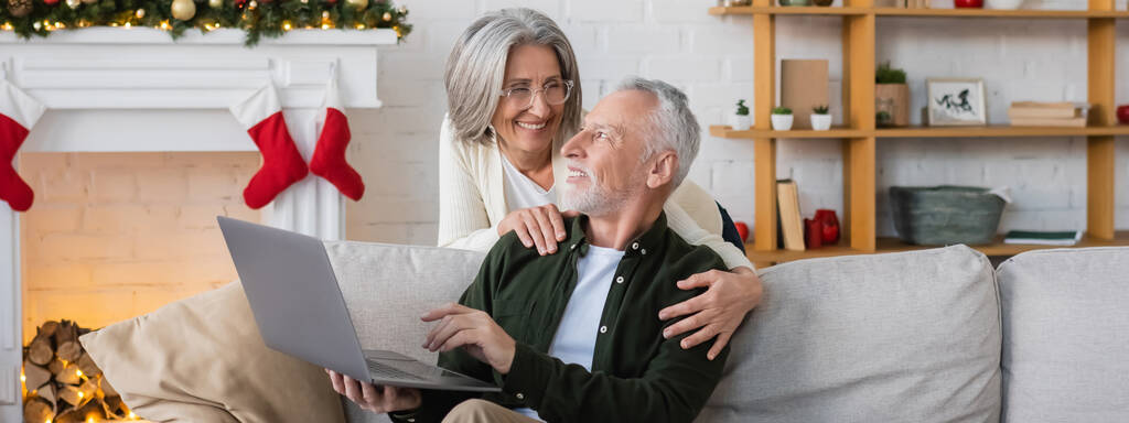 χαρούμενη μεσήλικη γυναίκα με γυαλιά αγκαλιάζει τον σύζυγο με το laptop την ημέρα των Χριστουγέννων, banner - Φωτογραφία, εικόνα