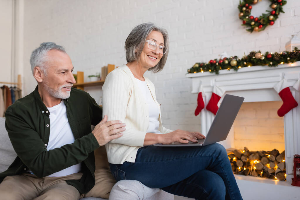 χαρούμενη μεσήλικη γυναίκα με γυαλιά χρησιμοποιώντας φορητό υπολογιστή κοντά χαρούμενος σύζυγος κατά τη διάρκεια των Χριστουγέννων διακοπές  - Φωτογραφία, εικόνα