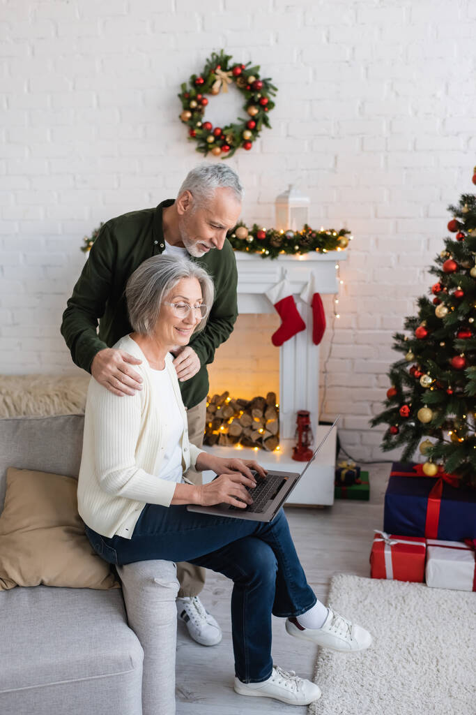 mężczyzna w średnim wieku przytulanie wesoły żona w okulary za pomocą laptopa w pobliżu choinki  - Zdjęcie, obraz