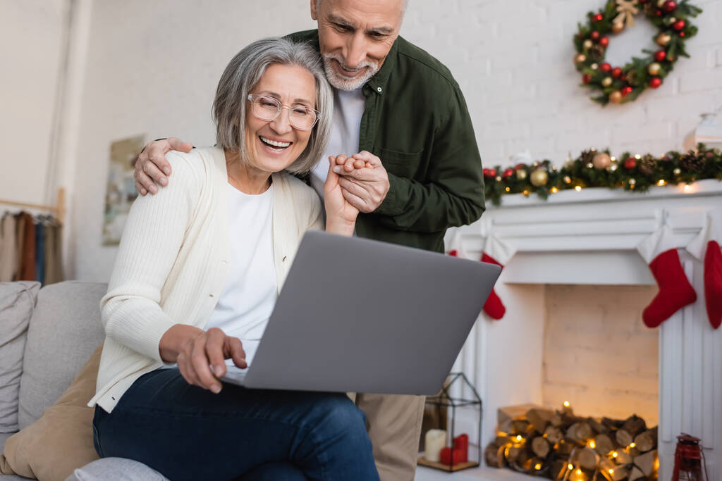 onnellinen keski-ikäinen nainen laseissa kannettavan tietokoneen avulla ja pitämällä kädestä miehensä kanssa joululomien aikana  - Valokuva, kuva