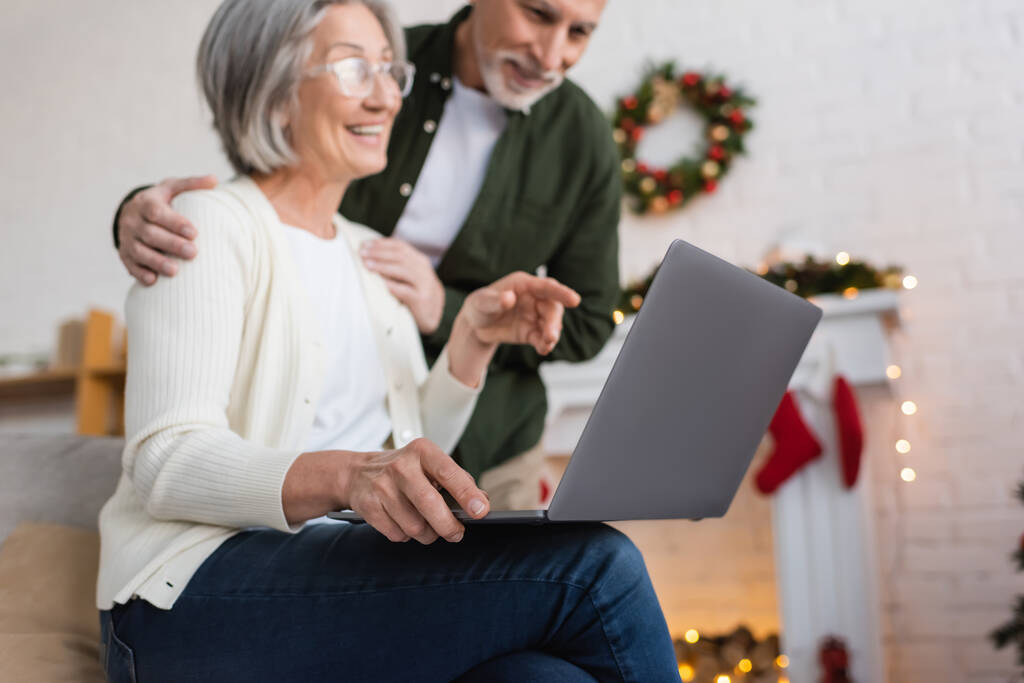 szczęśliwy w średnim wieku kobieta w okularach wskazując na laptopa w pobliżu szczęśliwy mąż podczas świąt Bożego Narodzenia  - Zdjęcie, obraz