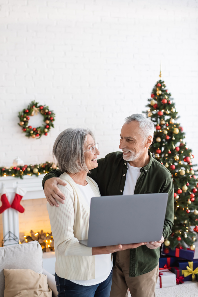 щасливий чоловік середнього віку обіймає дружину, тримаючи ноутбук біля прикрашеної ялинки
 - Фото, зображення