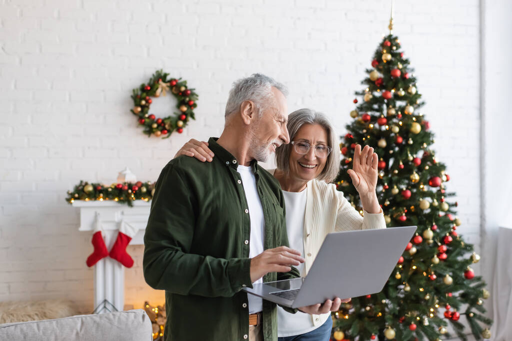 fröhlicher Mann mittleren Alters sieht Frau beim Videochat in der Nähe geschmückten Weihnachtsbaums winkend an - Foto, Bild