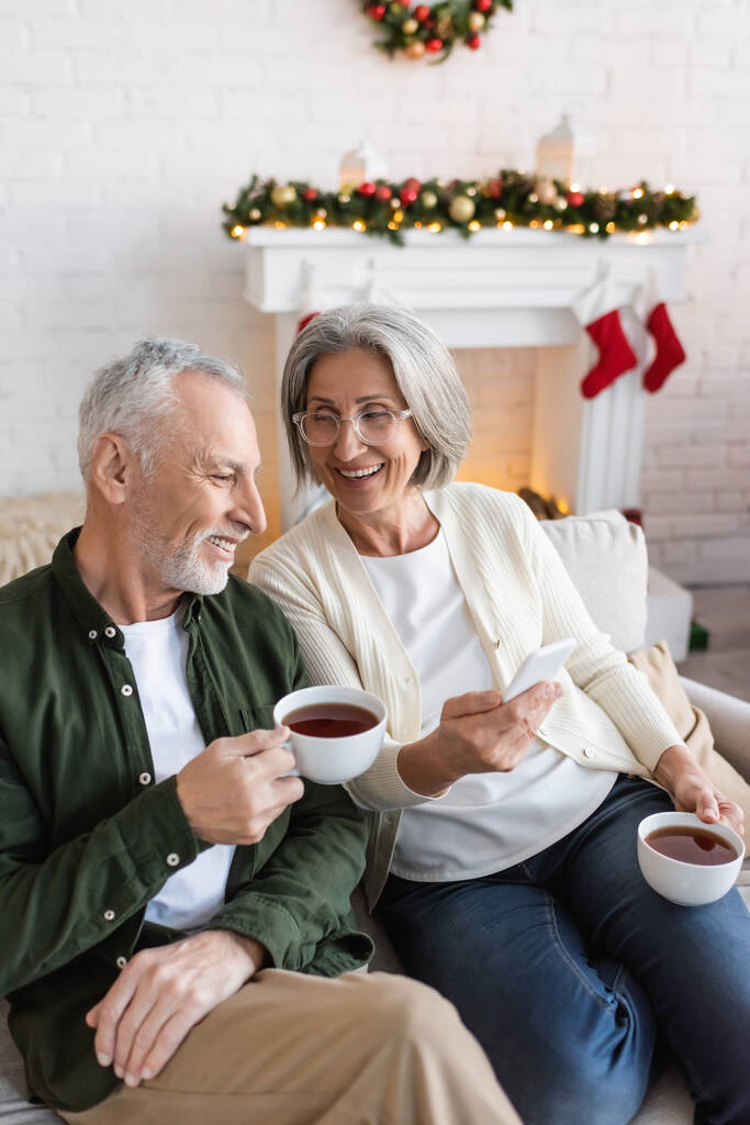 vrolijke en volwassen vrouw in bril houden smartphone in de buurt van echtgenoot met kopje thee tijdens kerstvakantie  - Foto, afbeelding