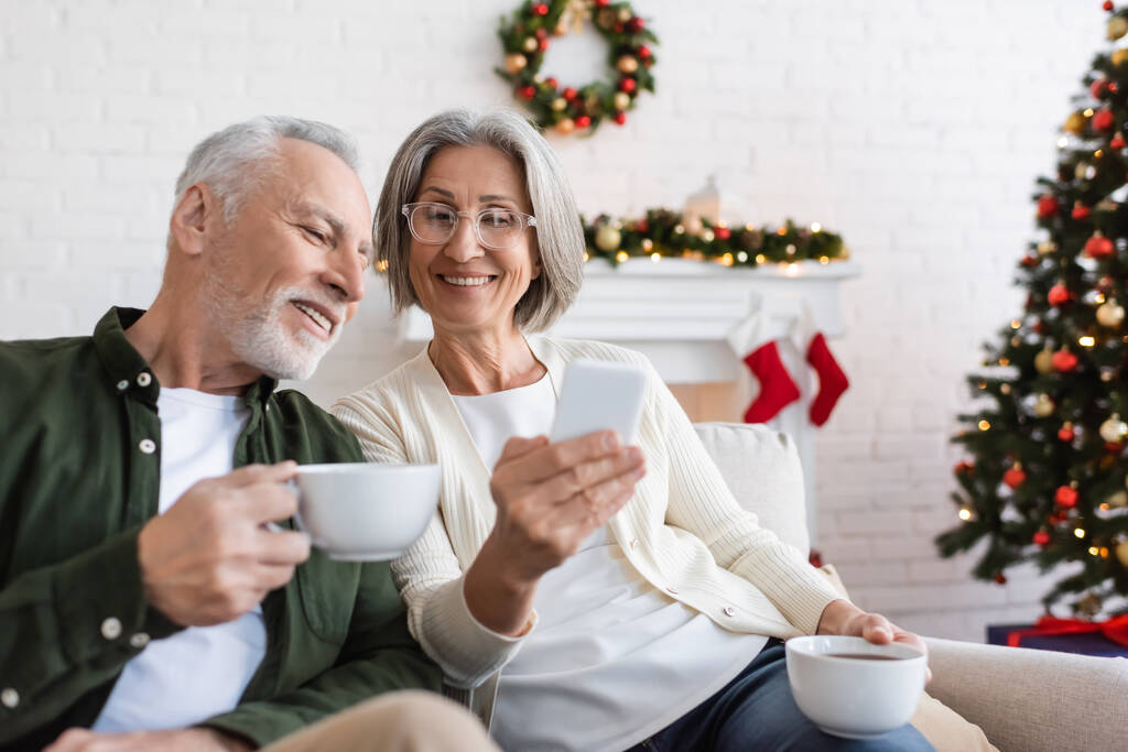 lächelnde und reife Frau in Gläsern, die während der Weihnachtsfeiertage ihr Smartphone in der Nähe ihres Mannes mit einer Tasse Tee hält  - Foto, Bild