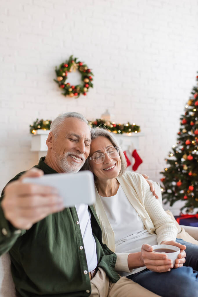 hombre de mediana edad tomando selfie con esposa feliz cerca del árbol de Navidad en casa - Foto, Imagen