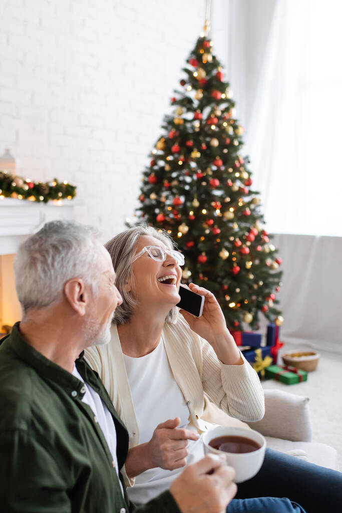 hombre maduro sosteniendo la taza de té y mirando a la esposa riendo mientras habla en el teléfono inteligente cerca del árbol de Navidad - Foto, imagen