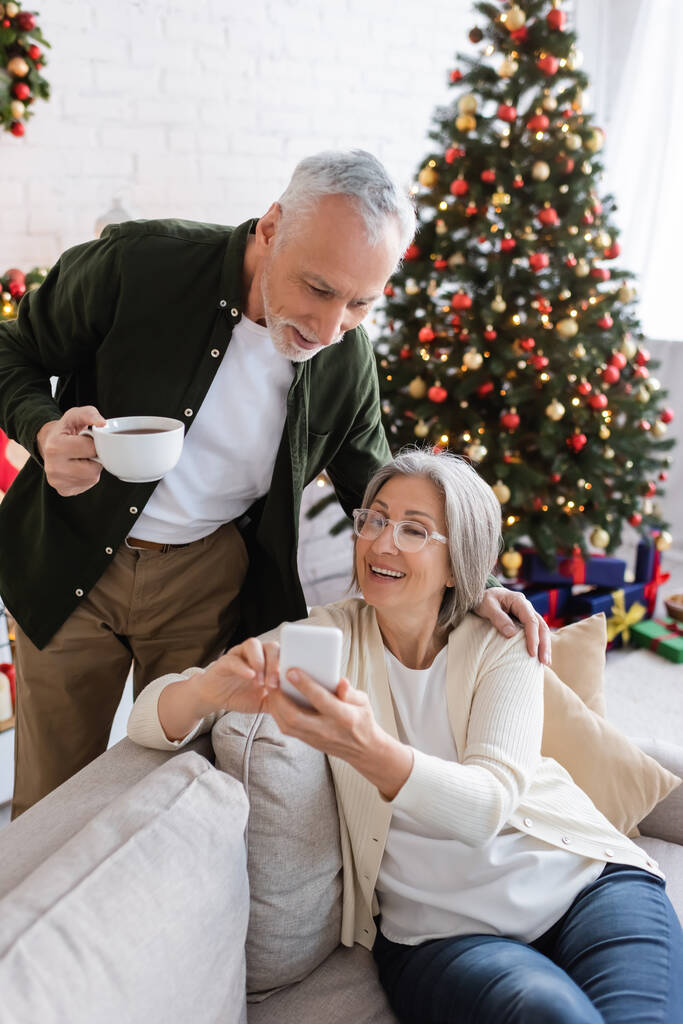 hombre maduro sosteniendo la taza de té y mirando a la esposa usando el teléfono inteligente cerca del árbol de Navidad - Foto, Imagen