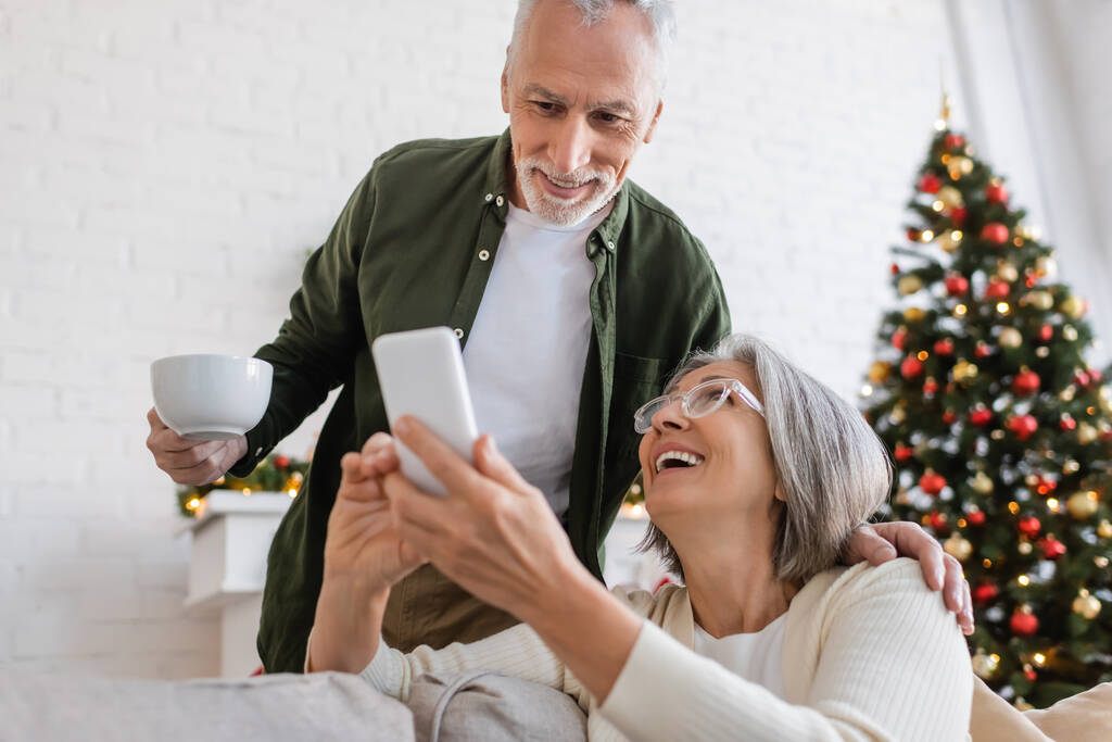 hombre maduro sosteniendo la taza de té y mirando el teléfono inteligente cerca de la esposa y el árbol de Navidad borrosa - Foto, imagen