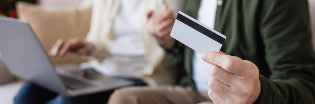 przycięty widok dojrzały mężczyzna posiadający kartę kredytową w pobliżu żony podczas dokonywania zakupów online na rozmytym tle, baner - Zdjęcie, obraz