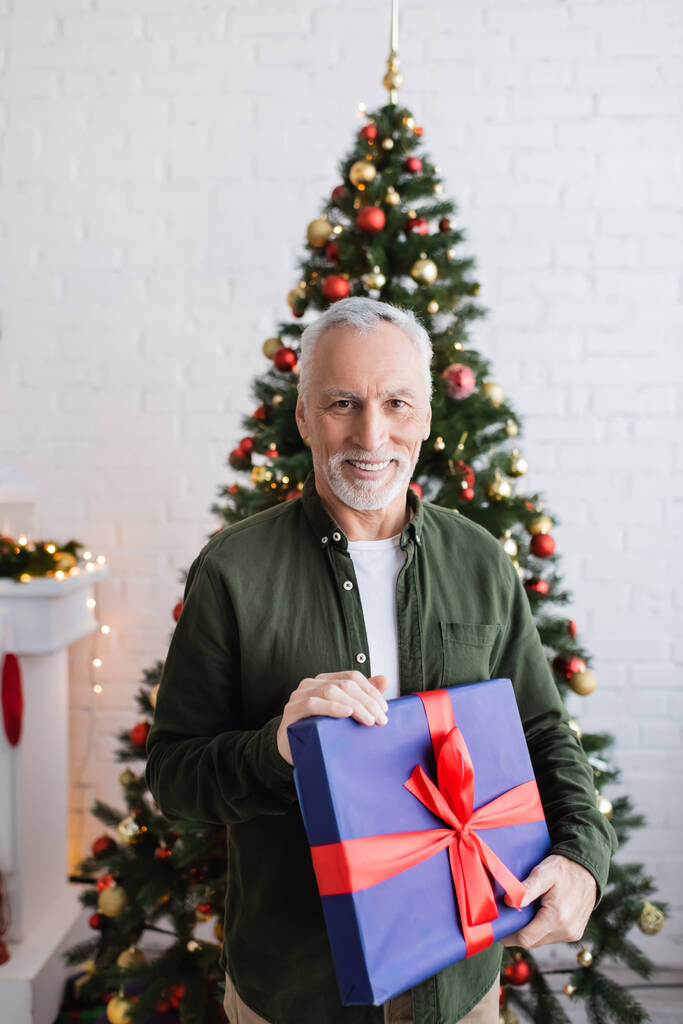 счастливый и бородатый мужчина средних лет держит завернутый подарок возле рождественской елки  - Фото, изображение