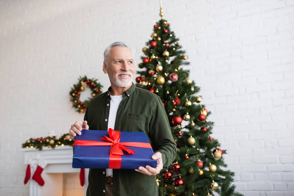 весёлый и бородатый мужчина средних лет держит завернутый подарок возле рождественской елки  - Фото, изображение