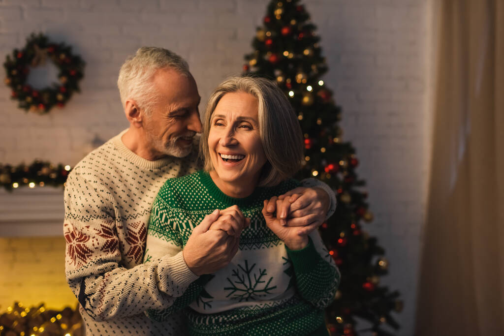 joyful bearded man in festive sweater hugging pleased mature wife near christmas tree in evening - Photo, Image