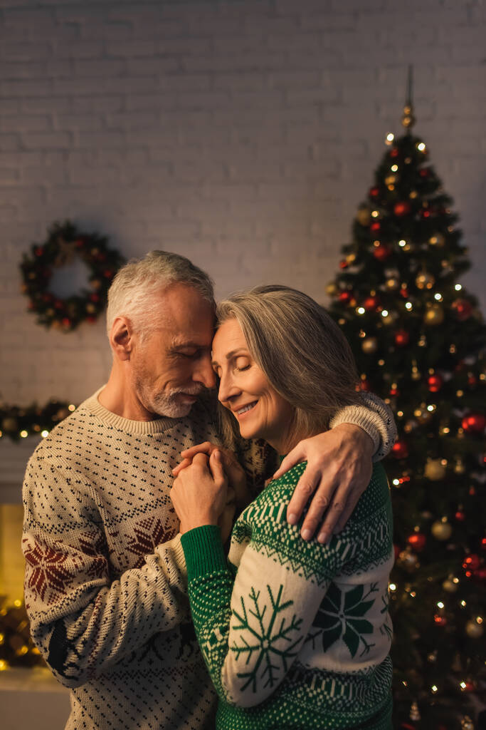 cheerful mature man in festive sweater hugging pleased wife near christmas tree in evening - Photo, Image