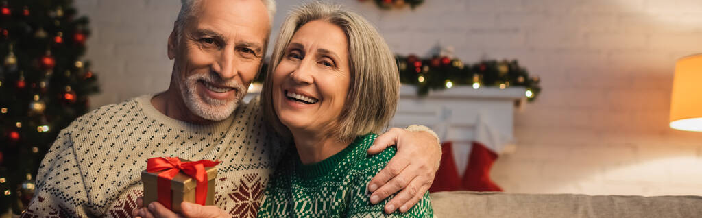 felice uomo di mezza età in maglione abbracciando moglie allegra con regalo di Natale, banner - Foto, immagini