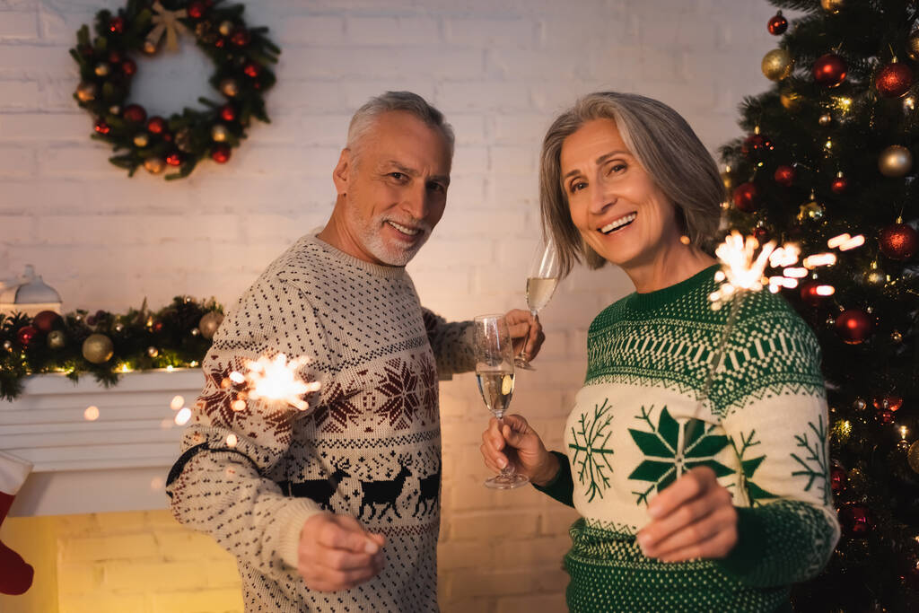 cheerful middle aged couple in festive sweaters holding bright sparklers and glasses of champagne on christmas eve - Photo, Image