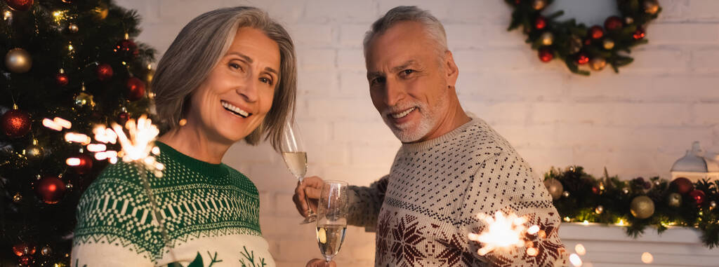 alegre pareja de mediana edad en suéteres festivos celebración de bengalas y copas de champán en la víspera de Navidad, bandera - Foto, imagen