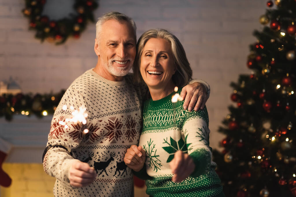 happy mature couple in holiday sweaters holding shiny sparklers on christmas eve at home - Photo, Image