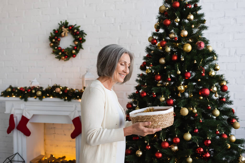 Neşeli ve olgun bir kadın elinde hasır sepetle evde süslü Noel ağacının yanında - Fotoğraf, Görsel