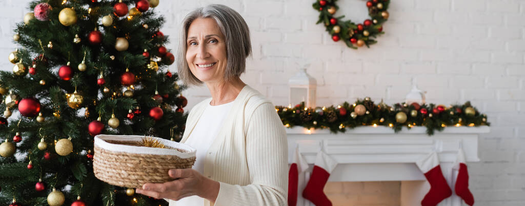 iloinen ja kypsä nainen tilalla paju kori lähellä koristeltu joulukuusi kotona, banneri - Valokuva, kuva