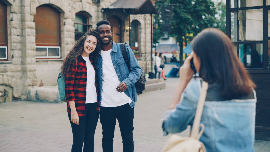Mladá žena fotí krásný pár Afroameričan a kavkazské dívky turisty stojící na ulici a pózující před kamerou. Cestovní ruch, technologie a přátelé koncept. - Fotografie, Obrázek