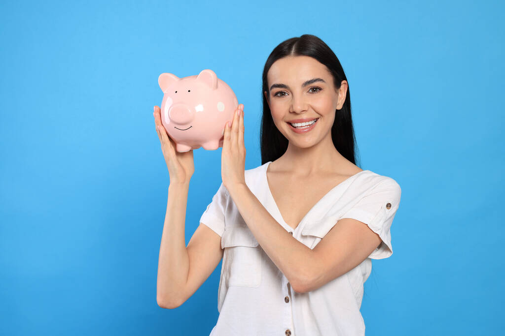 Glückliche junge Frau mit Sparschwein auf hellblauem Hintergrund - Foto, Bild